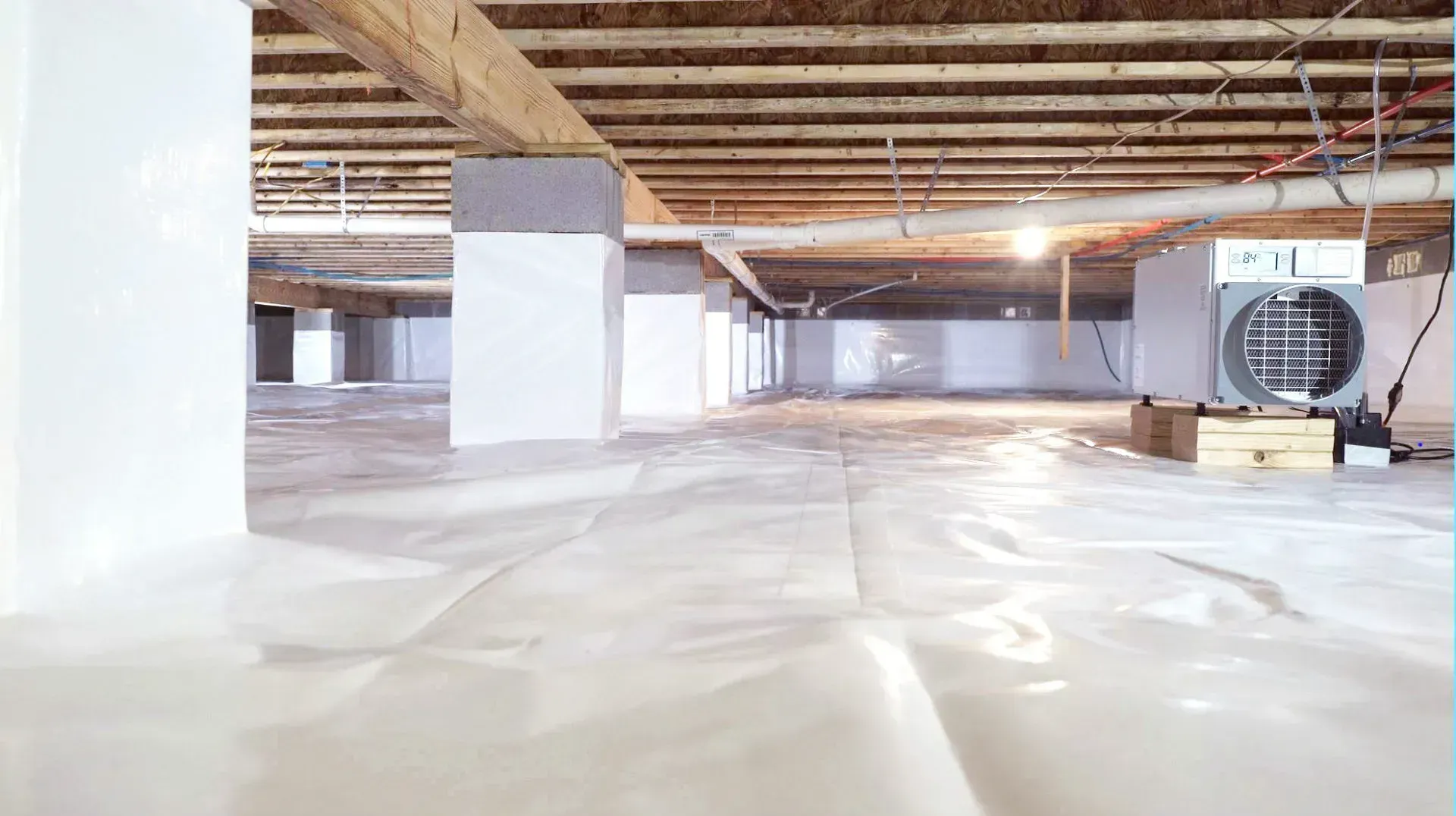 Brightly lit crawl space with white vapor barrier, supporting pillars, and exposed ceiling joists.