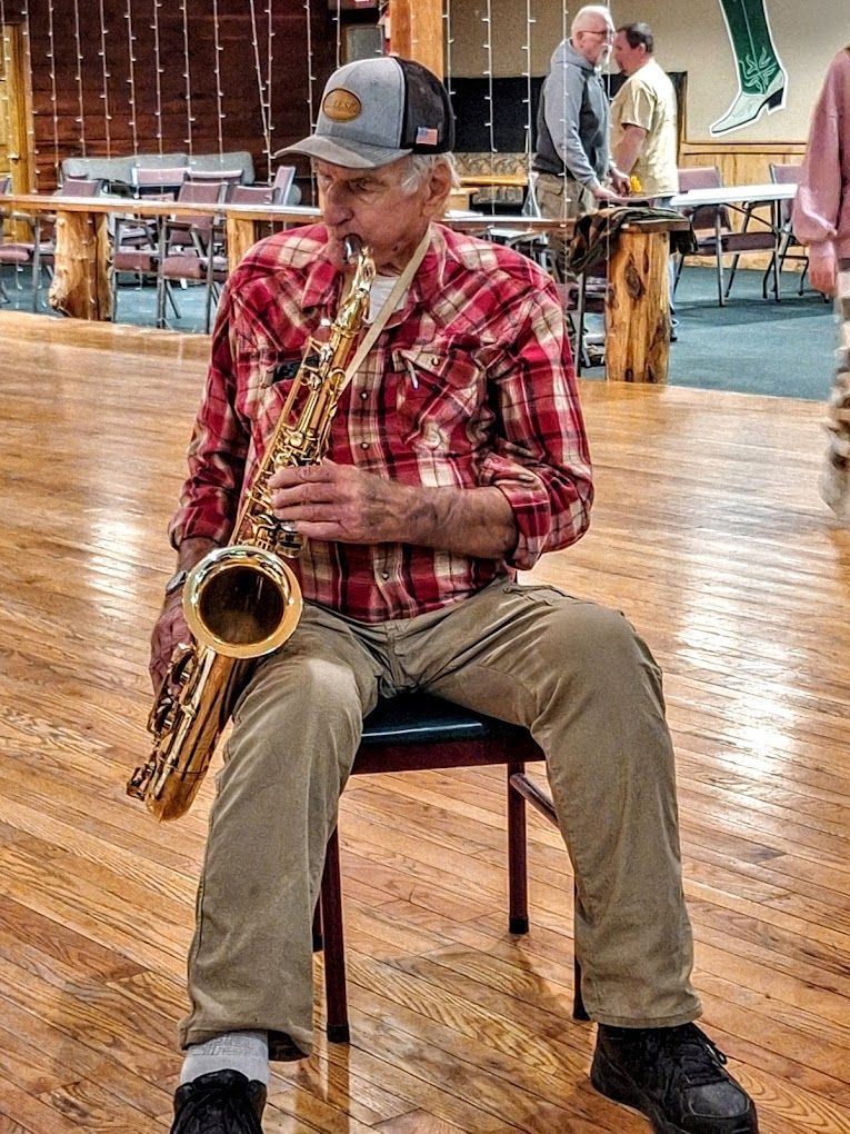 Man in red plaid shirt playing saxophone while seated in a wooden hall