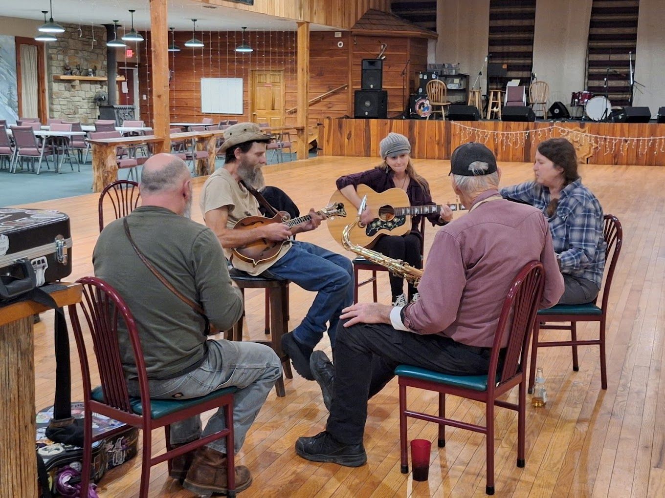 Five people sit in a rustic room playing guitars around a table with drinks and microphones.