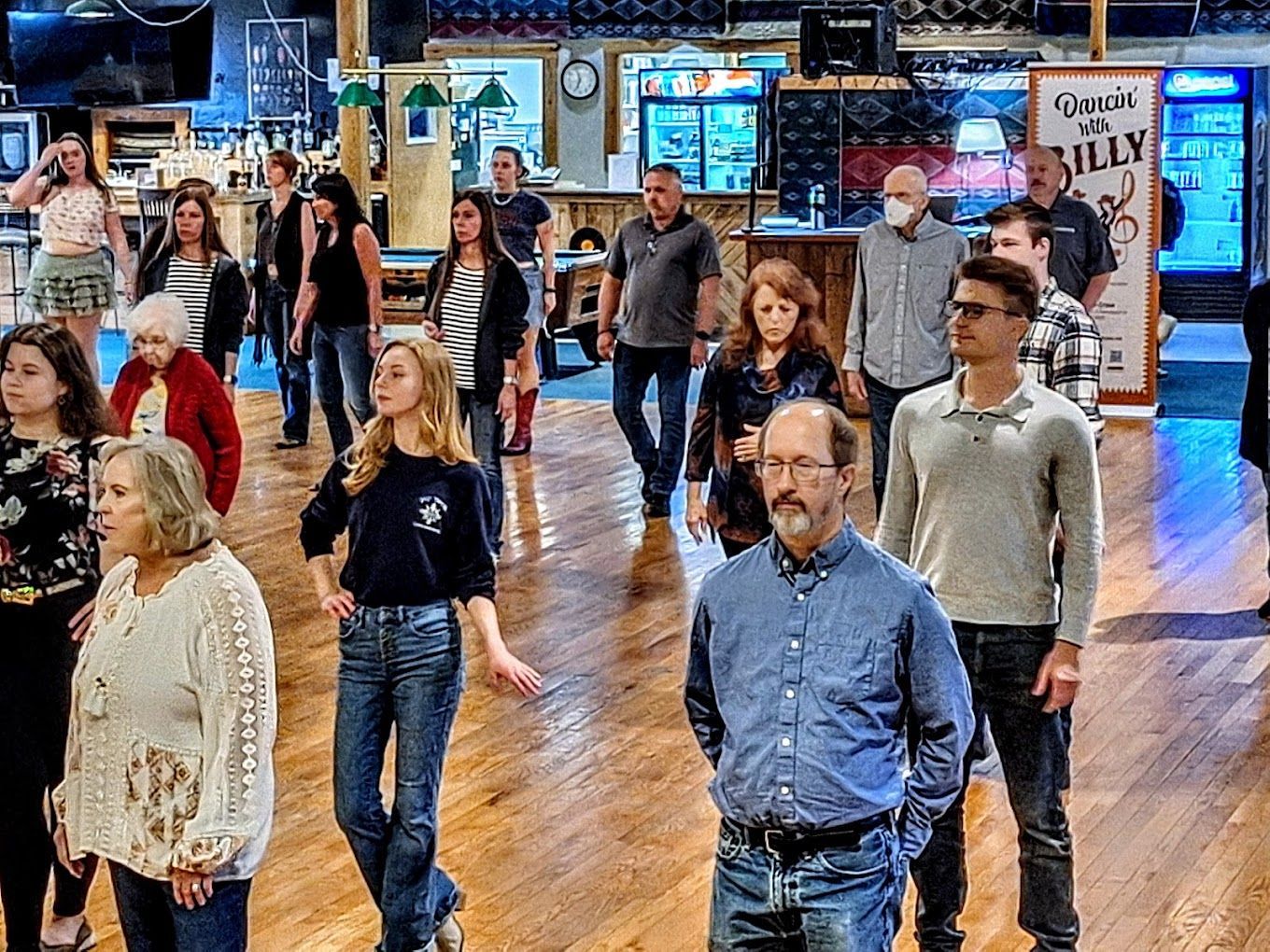 People standing and walking in a large indoor hall with a polished wooden floor and bright signs in the background
