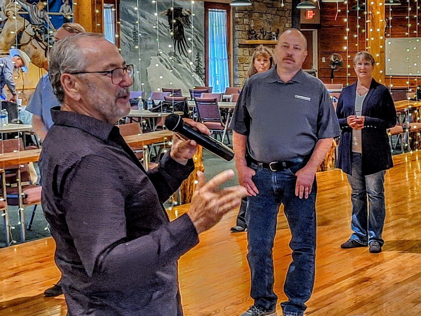 Man speaking to a small group in a bright lobby or showroom, with two people standing nearby holding drinks.