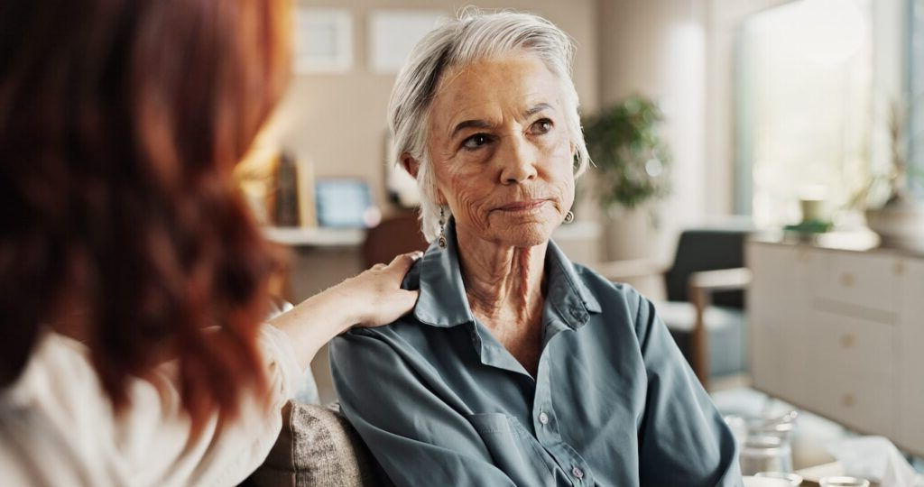 Two women talking in a bright living room; one listens with a serious expression.