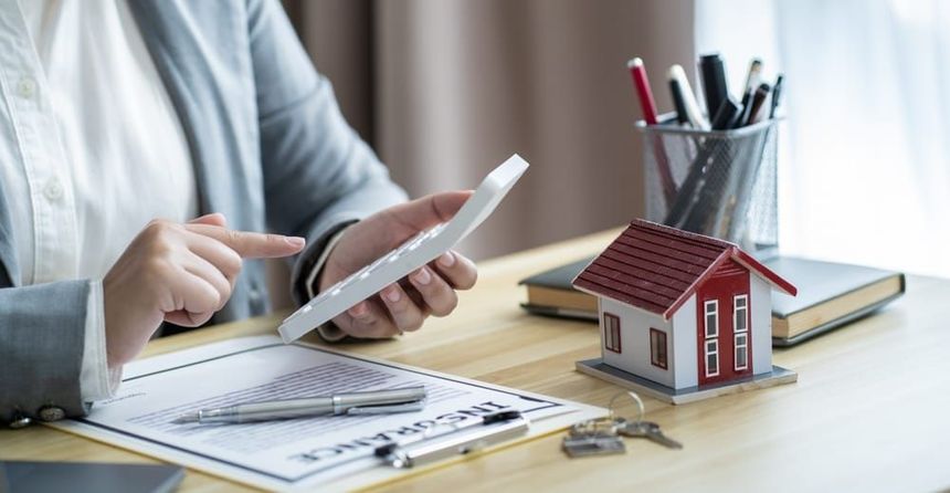 Person using a smartphone at a desk with house model, papers, calculator, and keys, suggesting real estate planning