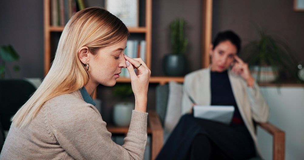 Woman in foreground wipes tears while another sits blurred in the background, suggesting a counseling session.