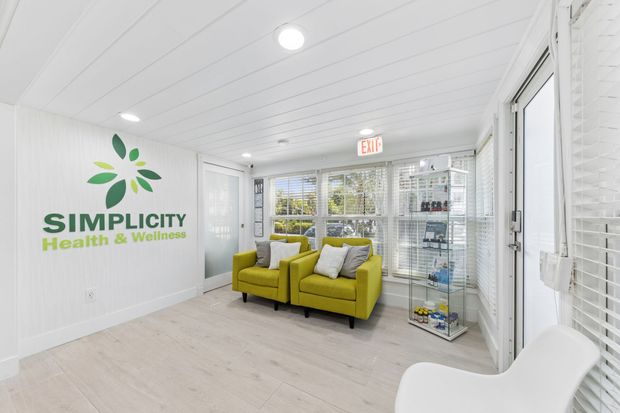 Bright white clinic waiting room with green sofa, white chair, and “Simplicity Health & Wellness” wall logo