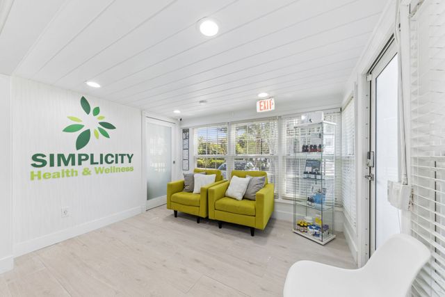 Bright white clinic waiting room with green sofa, white chair, and “Simplicity Health & Wellness” wall logo