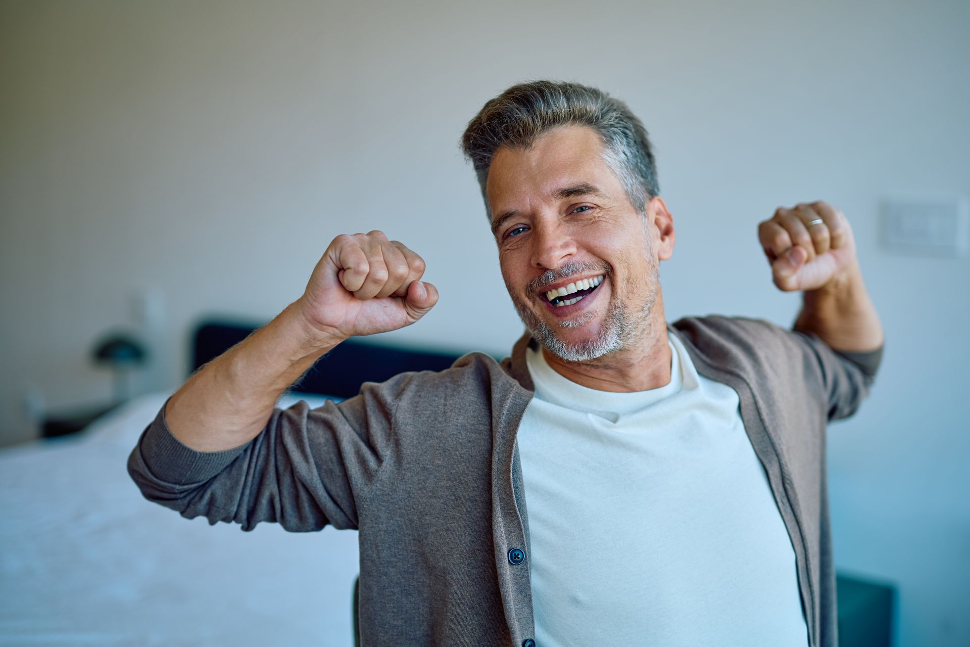A smiling person with grey hair wearing a cardigan stretches their arms up with fists clenched in a bedroom.
