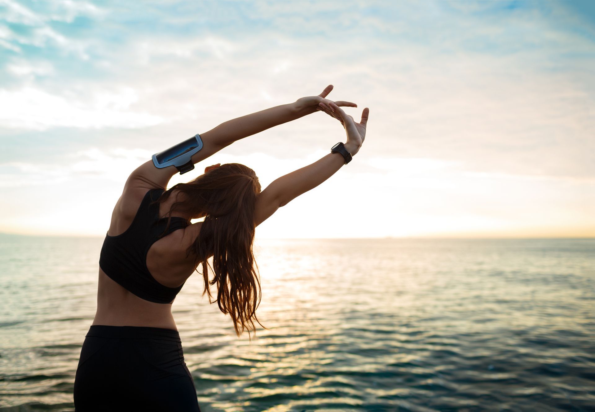 Woman stretching by the ocean at sunrise, wearing a black top with a fitness tracker.