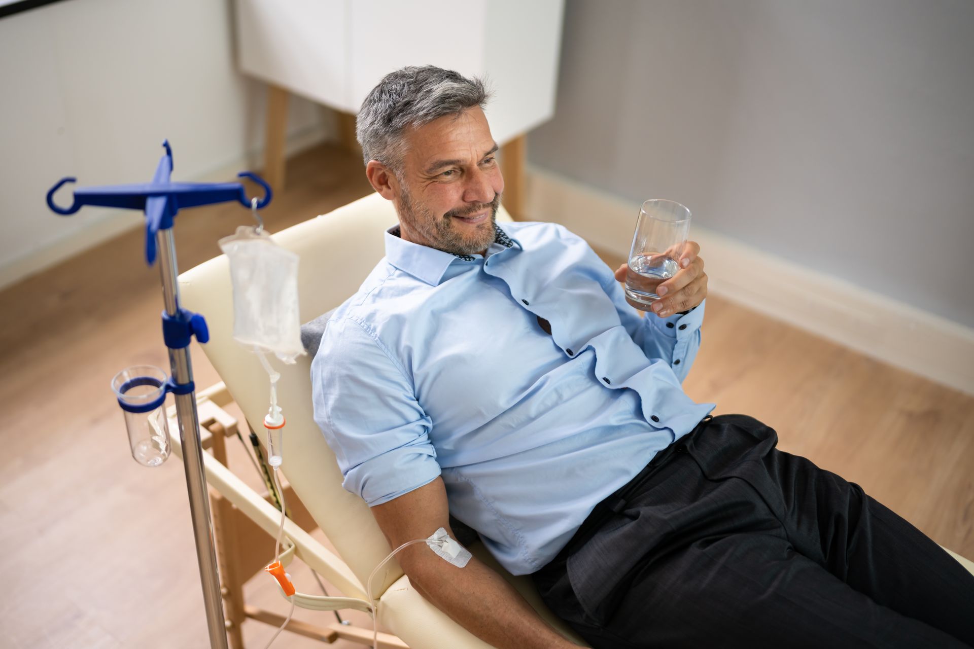 Man reclining in a chair receiving IV treatment while holding a glass, in a clinical setting