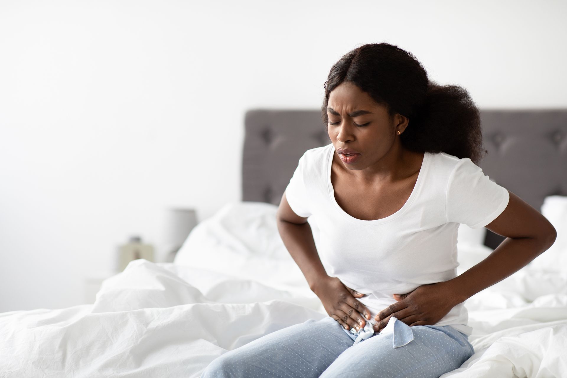 A person sitting on a bed, grimacing in pain while holding their lower abdomen with both hands.