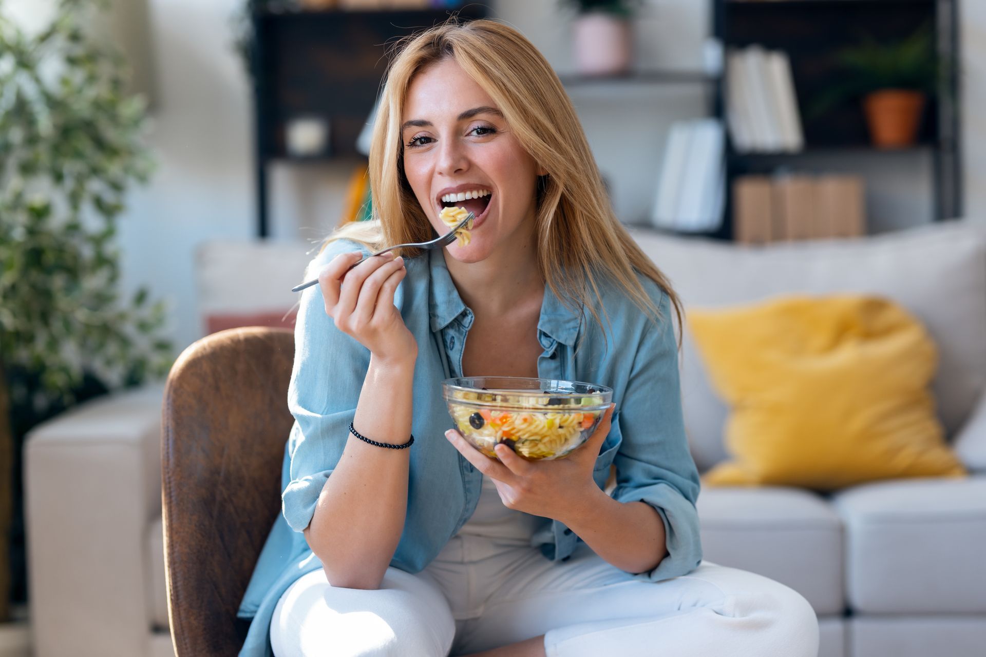 A person in a blue shirt smiling and eating a bowl of colorful fruit salad while sitting in a living room.