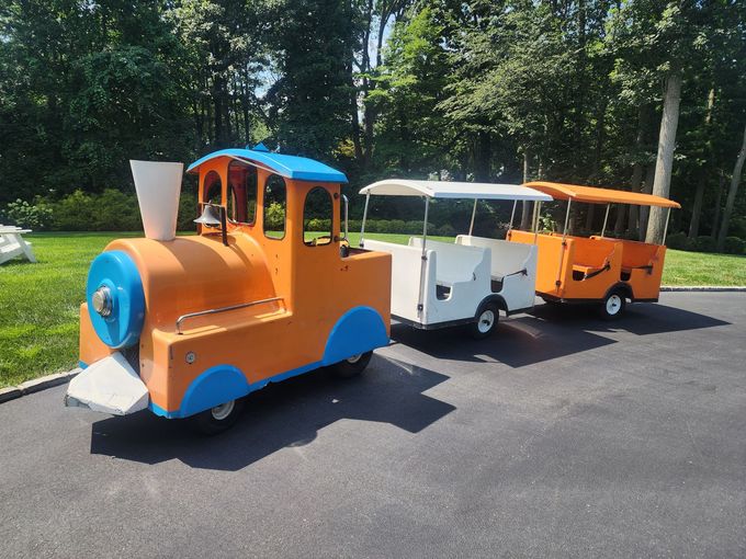 Red children's train ride with colorful carriages carrying passengers through a park on a sunny day