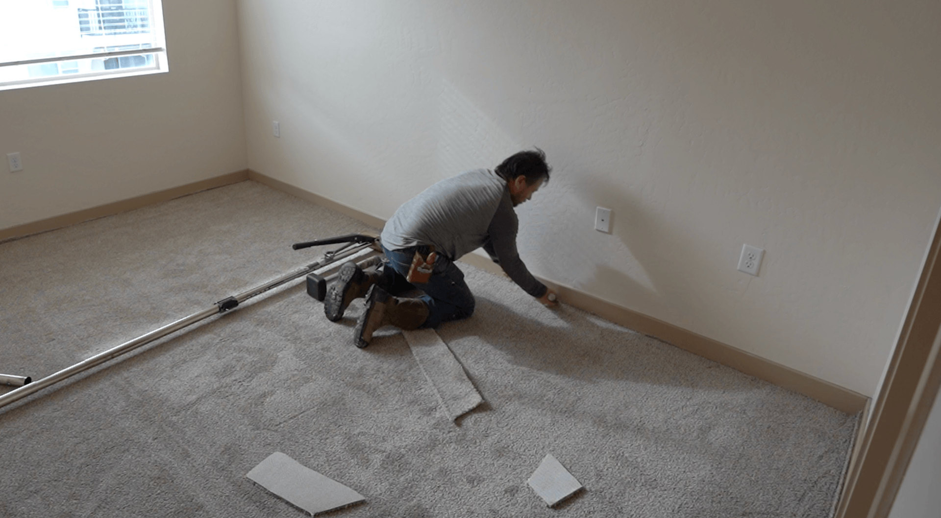 Man kneels on carpet in an empty room, smoothing flooring near the wall with tools beside him.