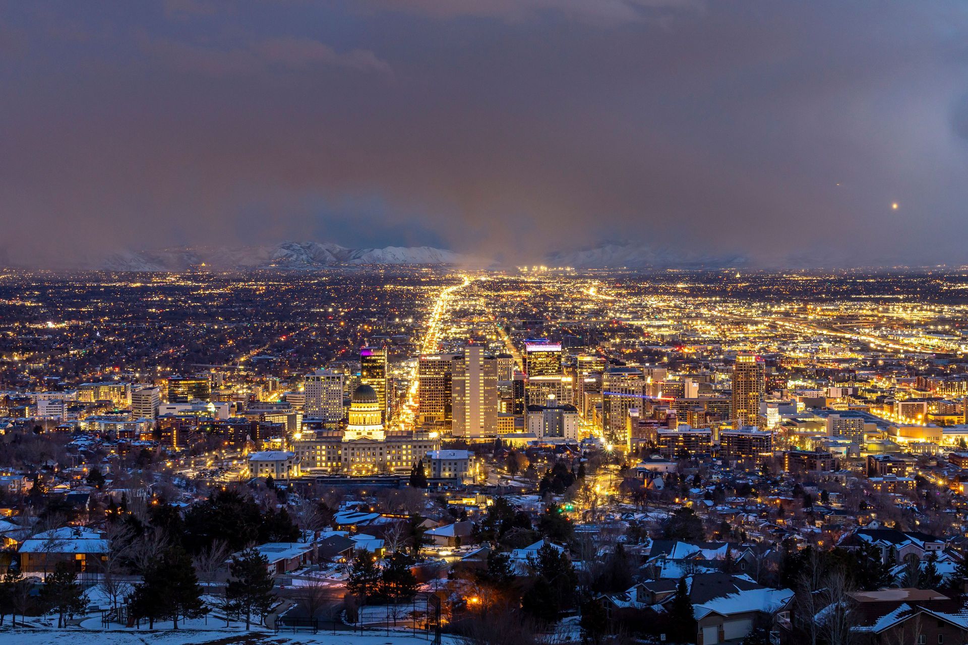 Snowy city skyline at dusk, glowing downtown lights beneath a purple evening sky.