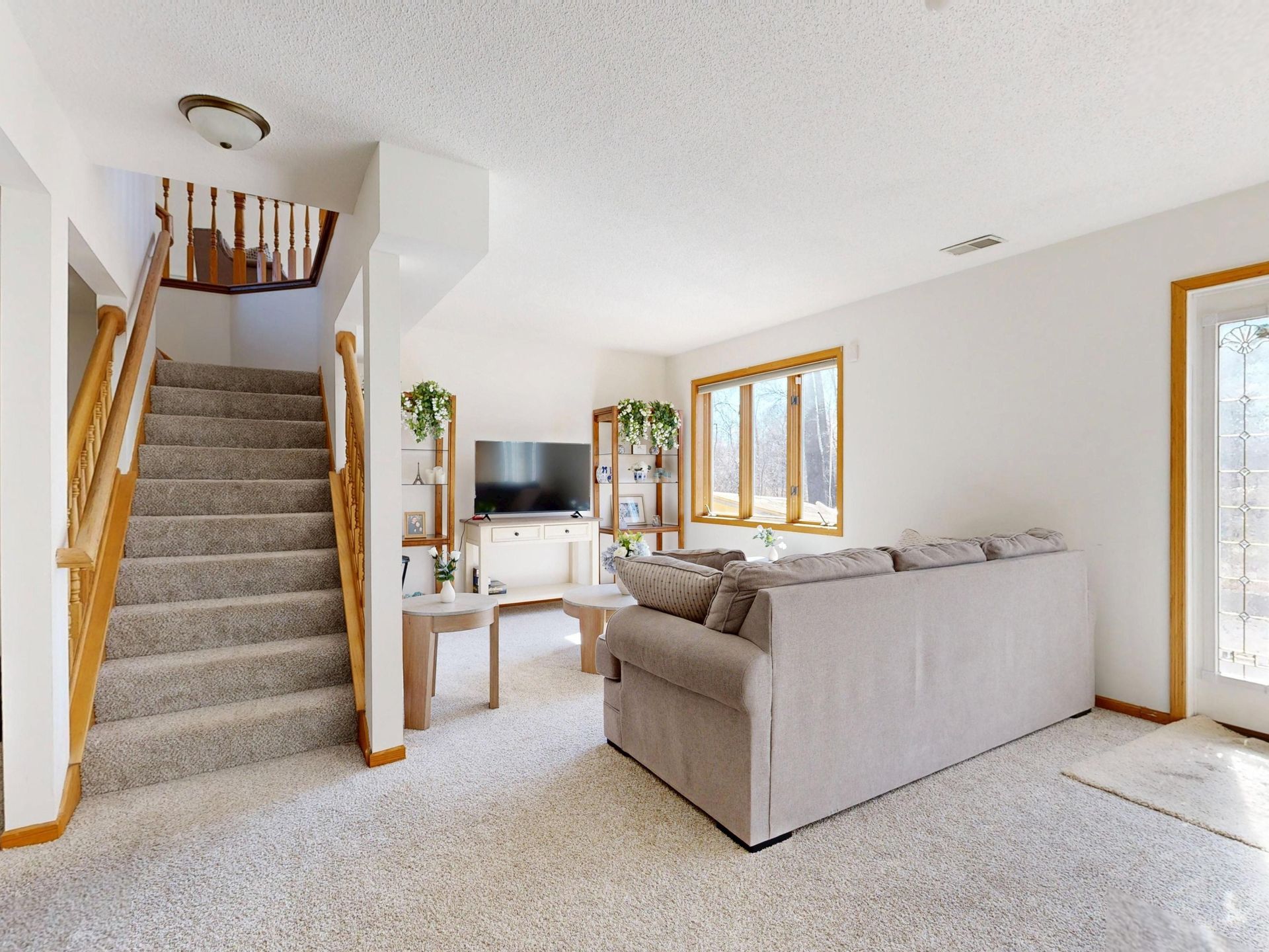 Carpeted living room with staircase, gray sofa, TV, and wood-trimmed windows and door.