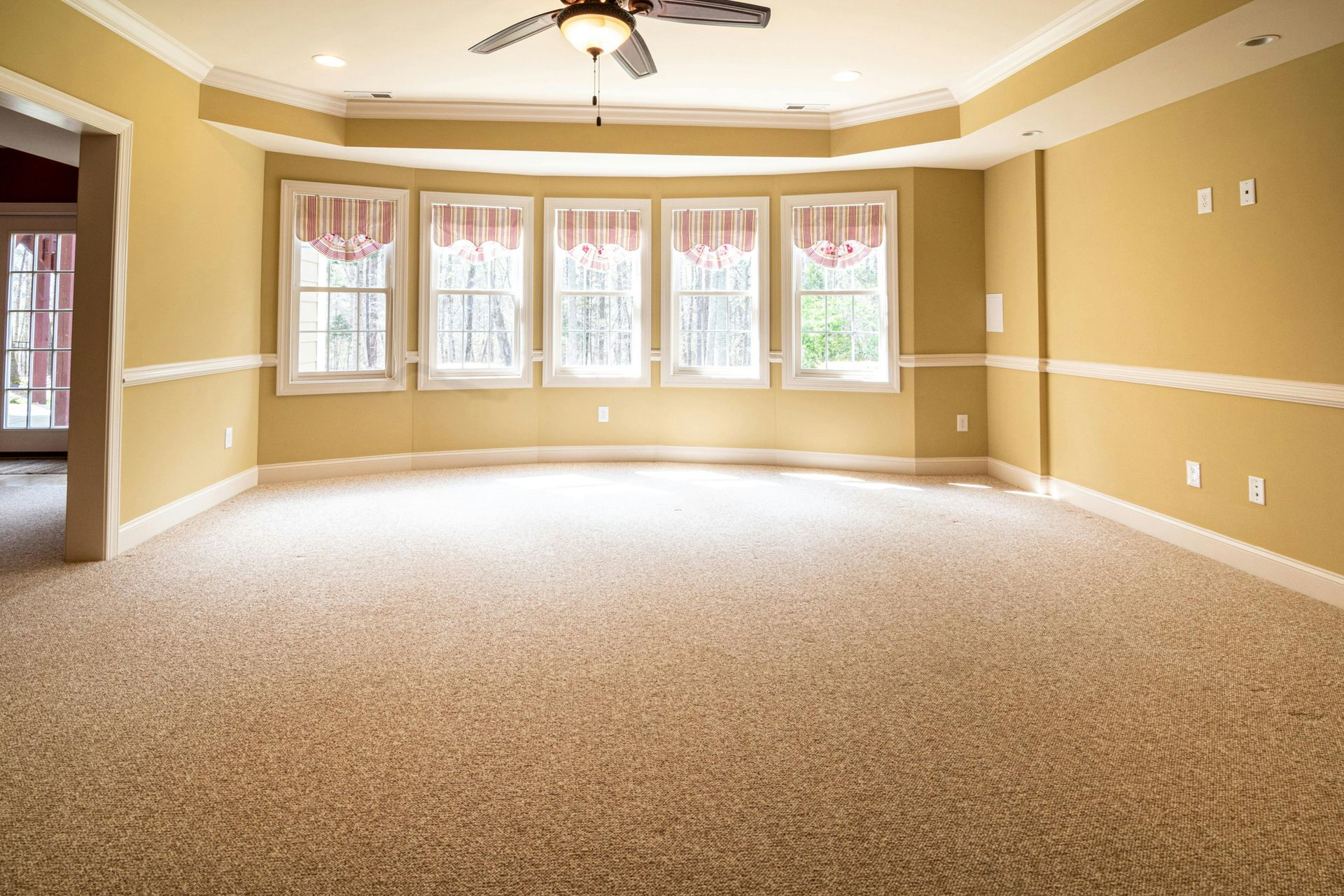Empty beige living room with carpet, tray ceiling fan, and large windows letting in bright daylight