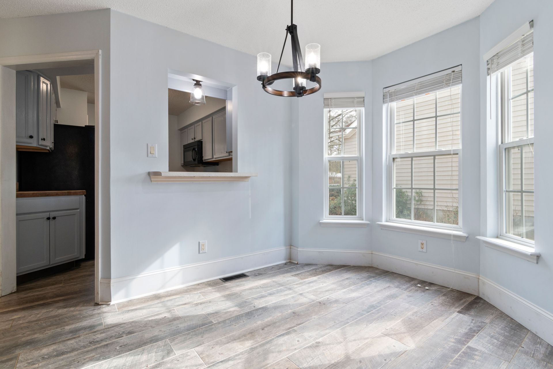Bright empty dining room with light blue walls, wood floors, bay windows, and a black chandelier
