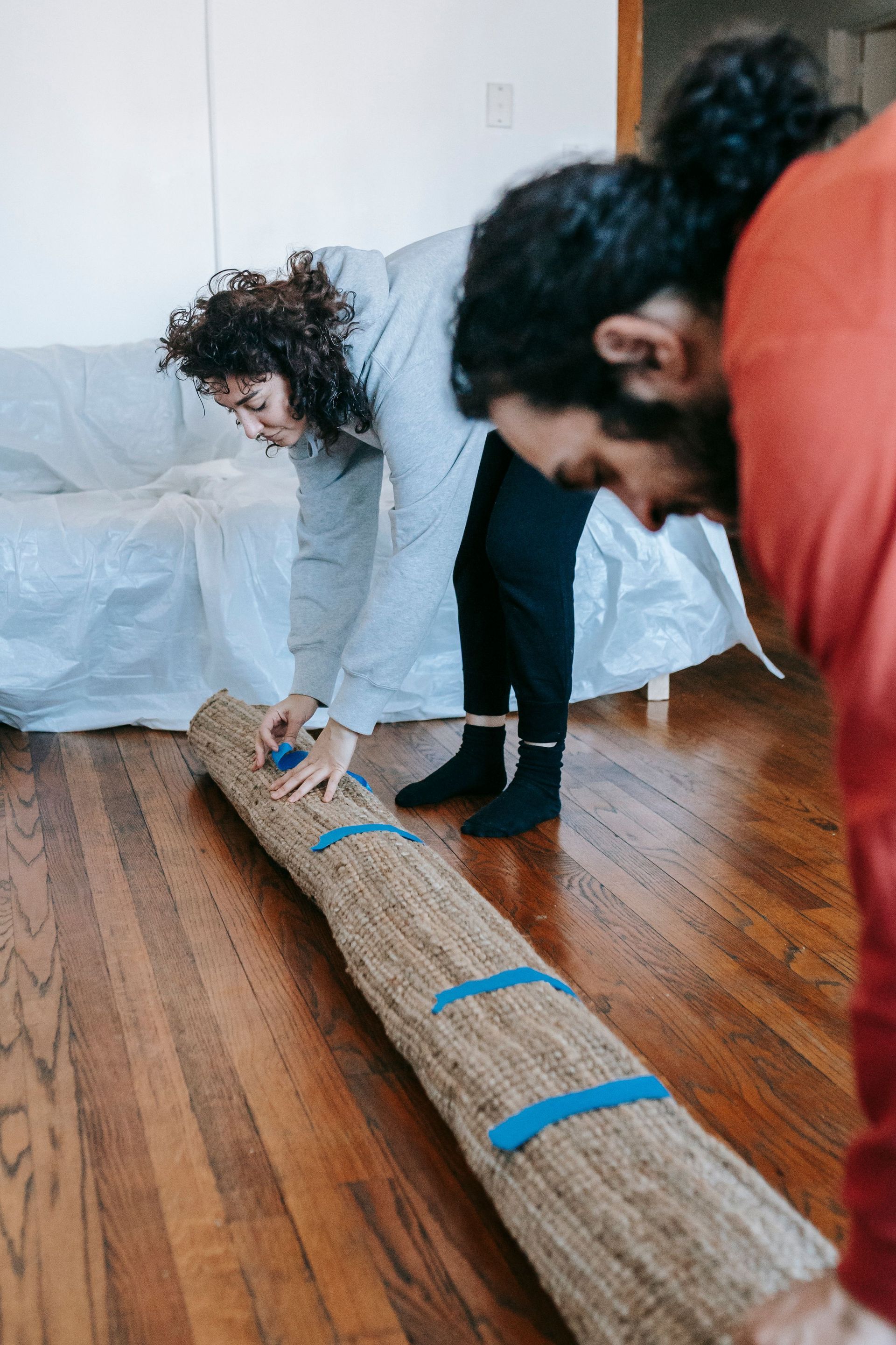 Two people securing a rolled straw mat on a hardwood floor with blue tape