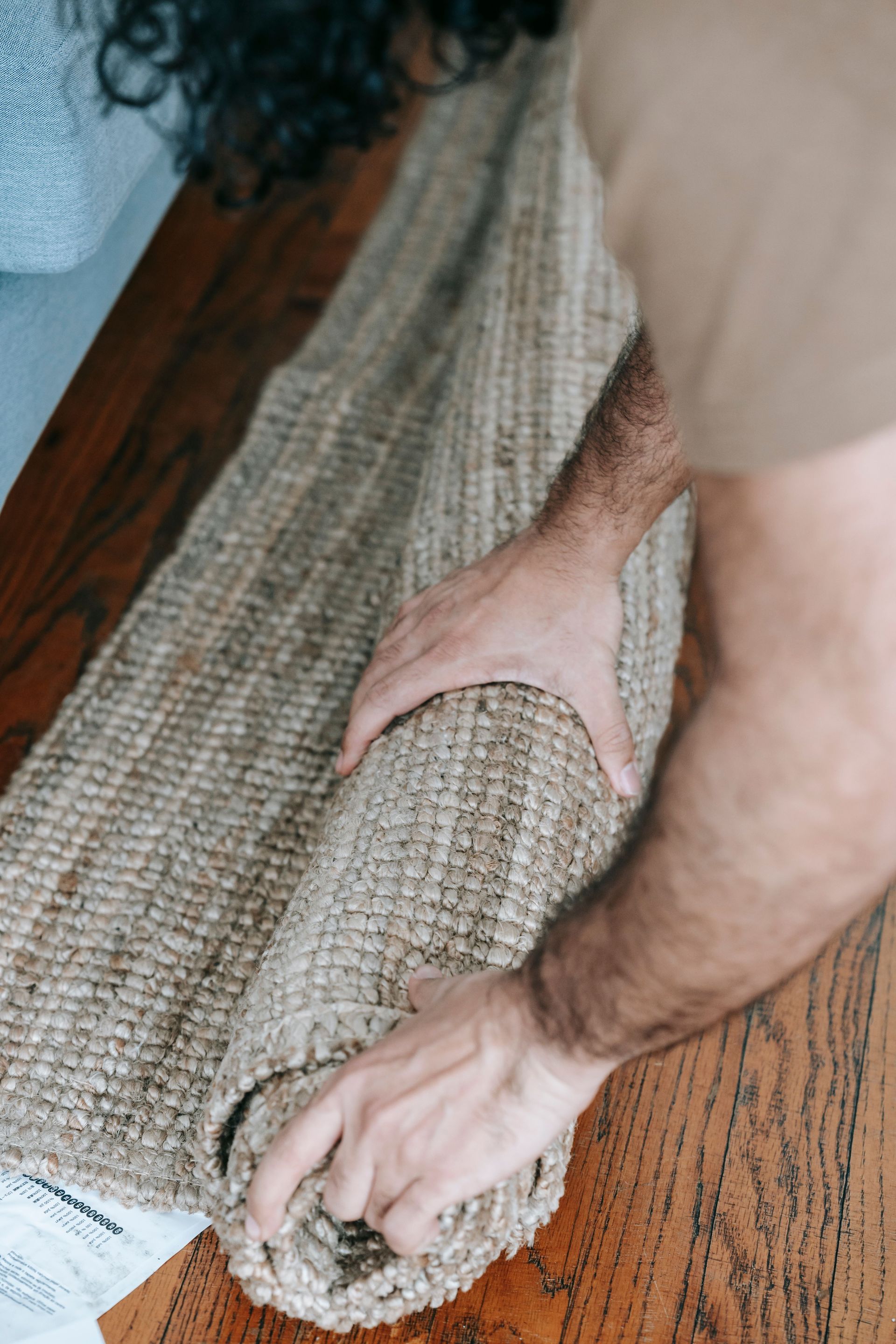 Hands rolling a woven beige rug on a wooden floor.