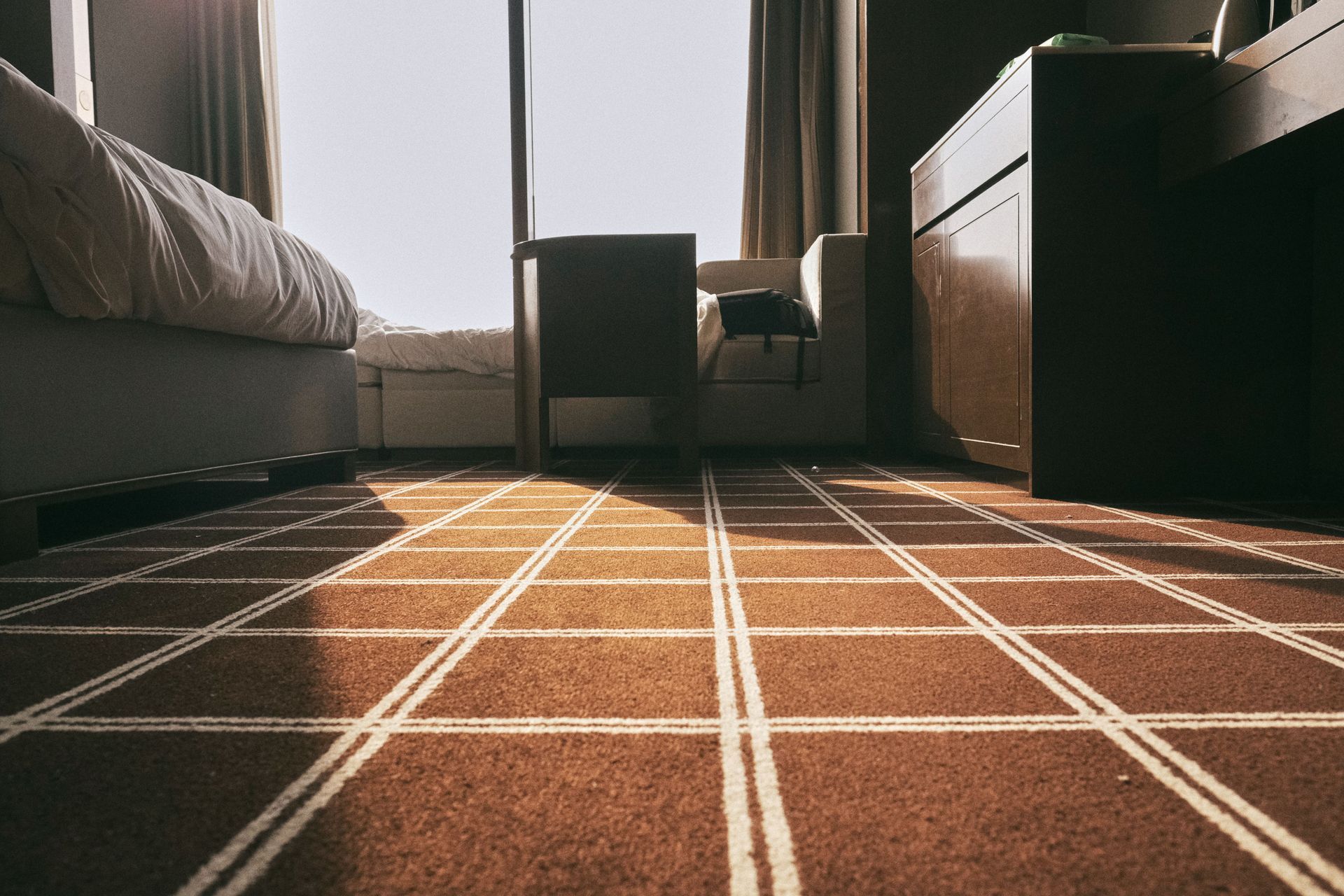 Low-angle view of a hotel room with a bed, chair, and sunlit window over a patterned carpet.