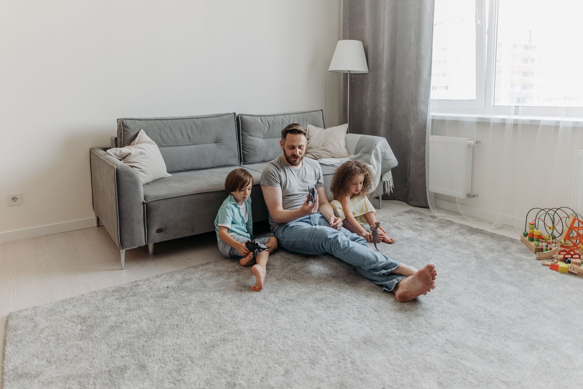 Family sitting on a gray couch in a bright living room, with two children playing on the carpet.