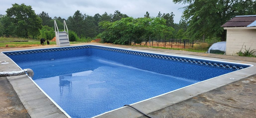 An empty rectangular swimming pool with blue tiles, surrounded by a concrete deck and a lush green landscape.