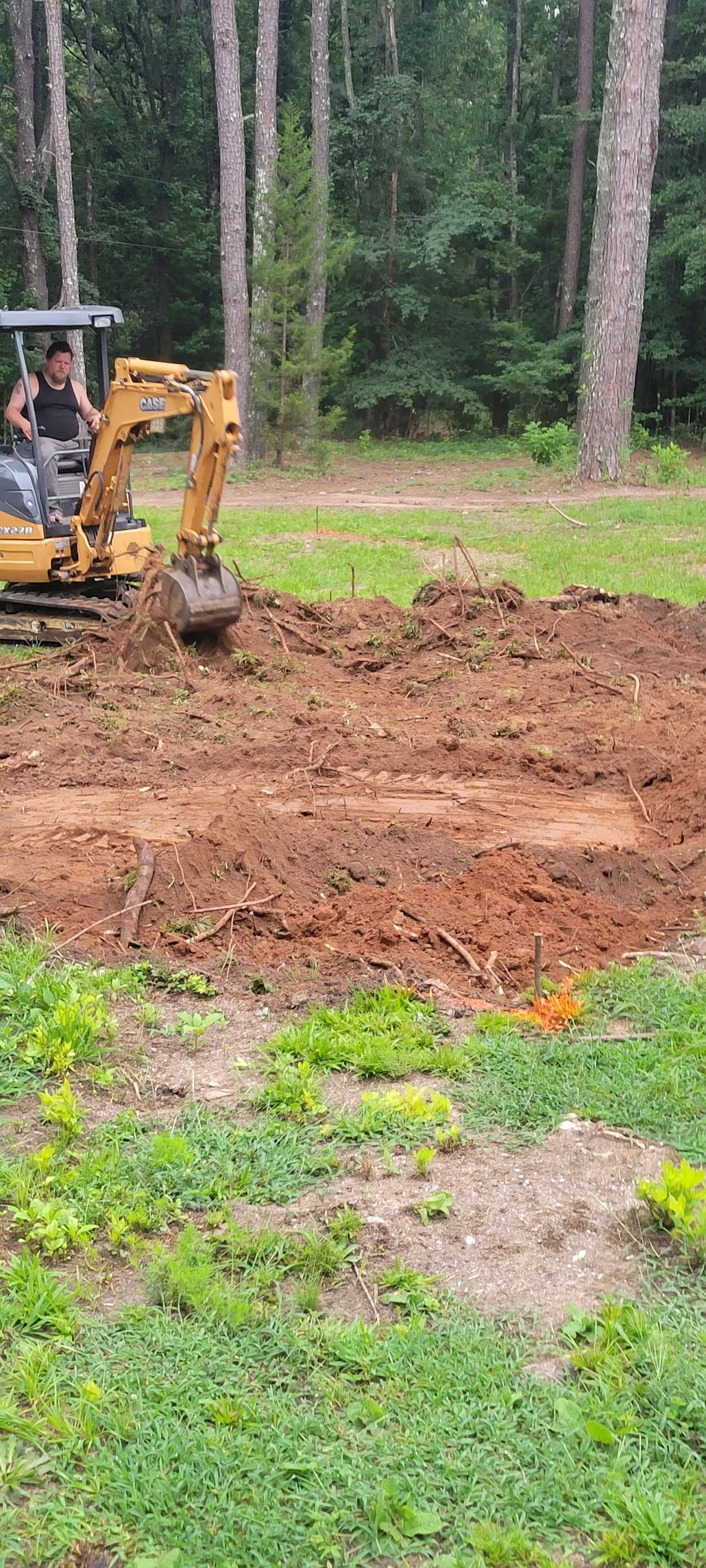 A person operates a yellow excavator, digging a hole in a dirt area surrounded by grass and trees.