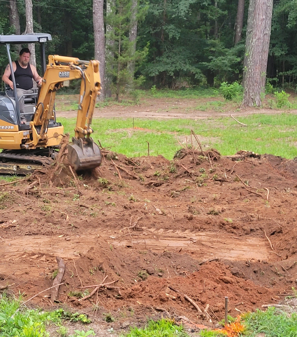 A person operating a yellow excavator, clearing dirt and debris in a wooded area.