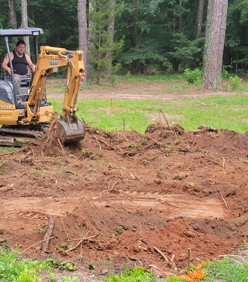 A person operating a yellow excavator, clearing dirt and debris in a wooded area.