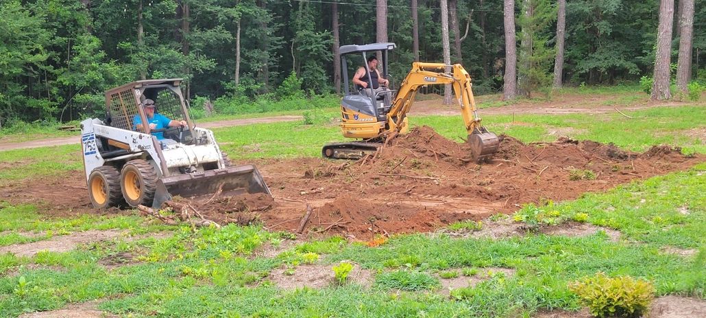 Two construction vehicles excavating dirt in a grassy area, trees in the background.