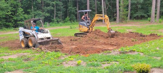 Two construction vehicles excavating dirt in a grassy area, trees in the background.