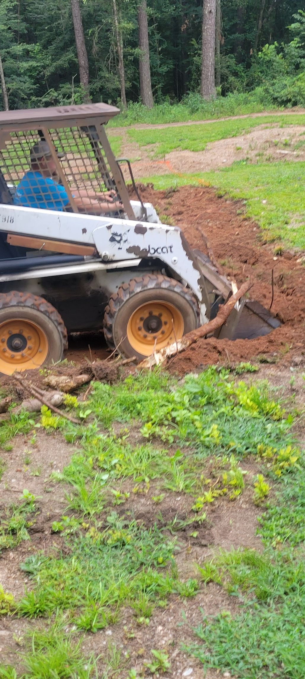 A man operating a Bobcat excavator digging a trench in a grassy area with trees in the background.
