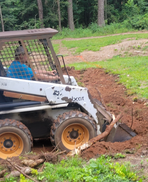 Person operating a white Bobcat digging in brown soil; green grass and trees in background.
