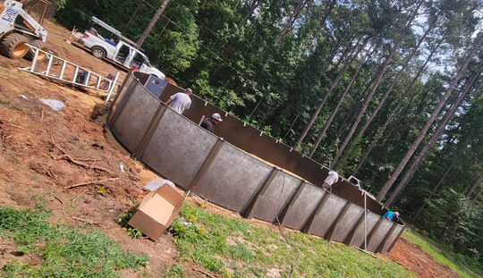 Workers assembling an above-ground pool on a sloped, dirt yard, with trees in the background.