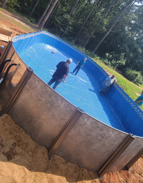 People in a newly-built above-ground pool. Brown siding, blue water, green grass, and trees in the background.
