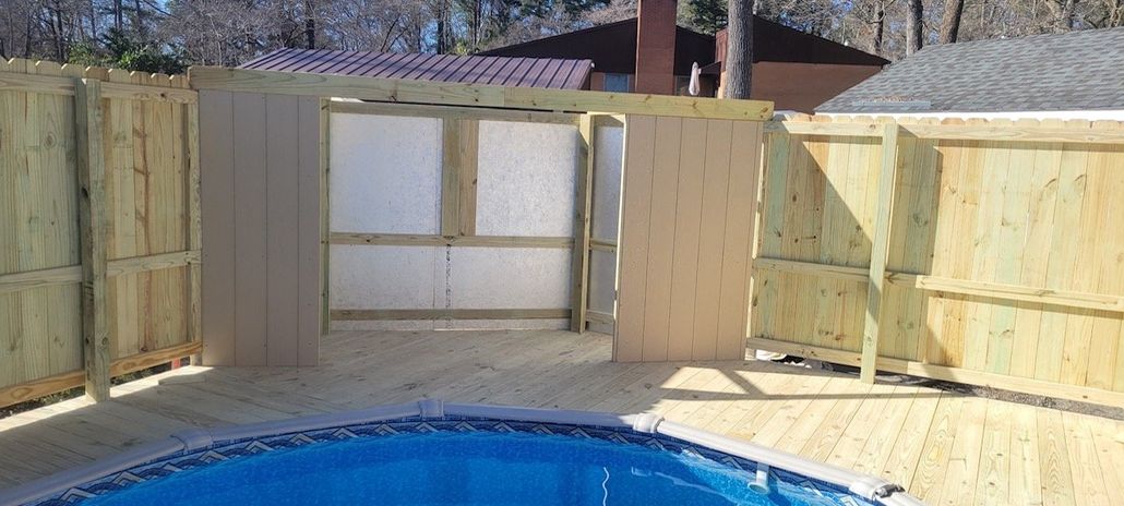 Wooden fence surrounds a pool with a deck. The fence has a gate that opens into a structure with white panels.