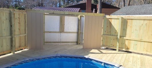 Wooden fence surrounds a pool with a deck. The fence has a gate that opens into a structure with white panels.