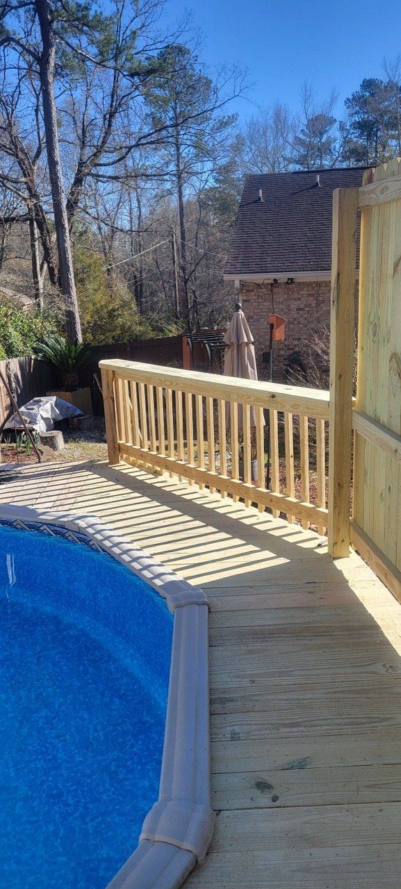 Wooden deck with a pool. A wooden railing runs along the pool. Trees and a house in the background.