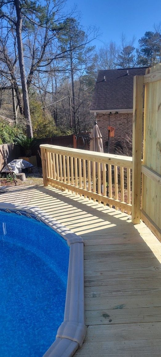 Wooden deck with a pool. A wooden railing runs along the pool. Trees and a house in the background.