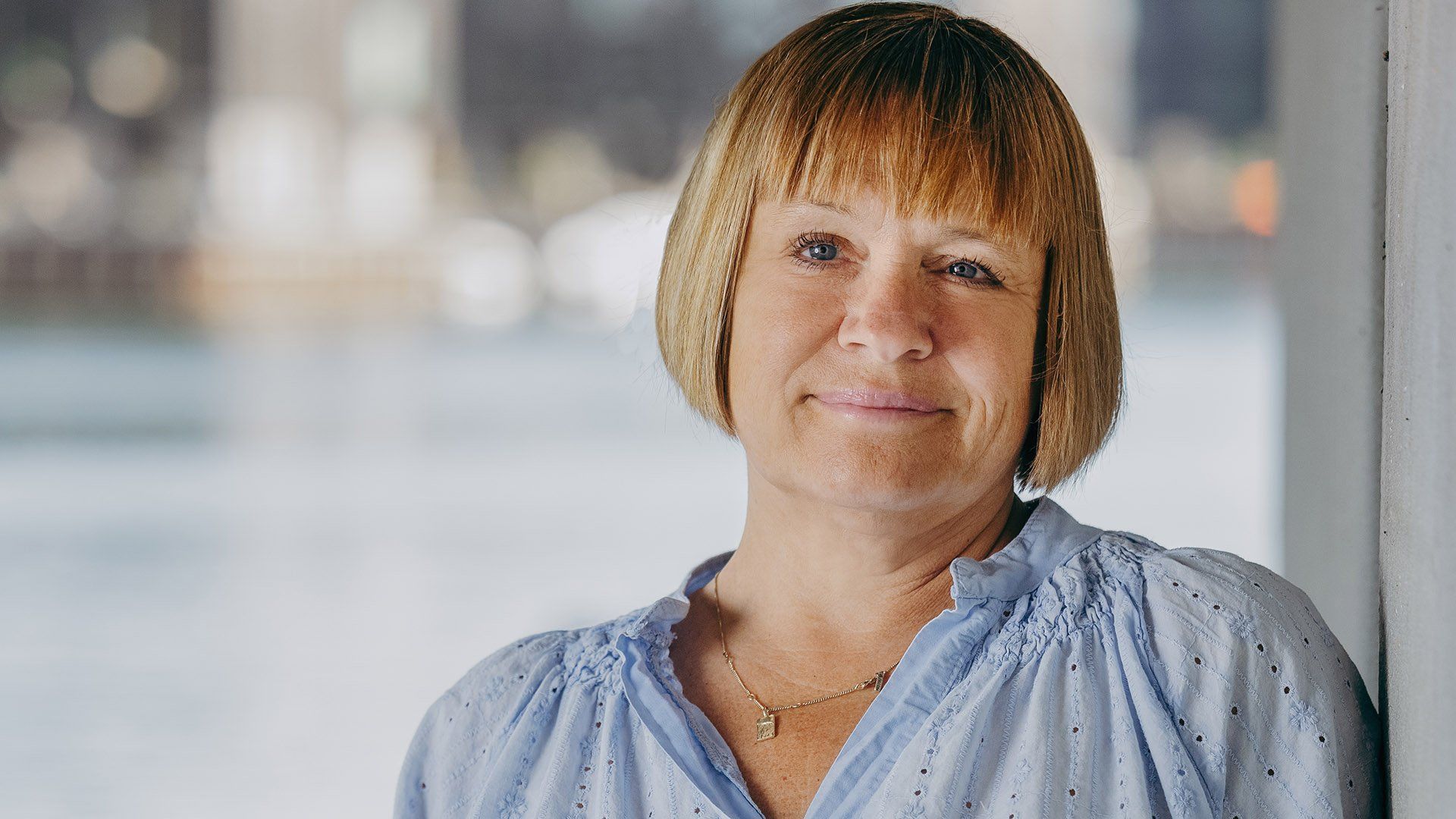 Kvinde med kort blond hår og blå bluse, smilende, lænet op ad en hvid væg.
