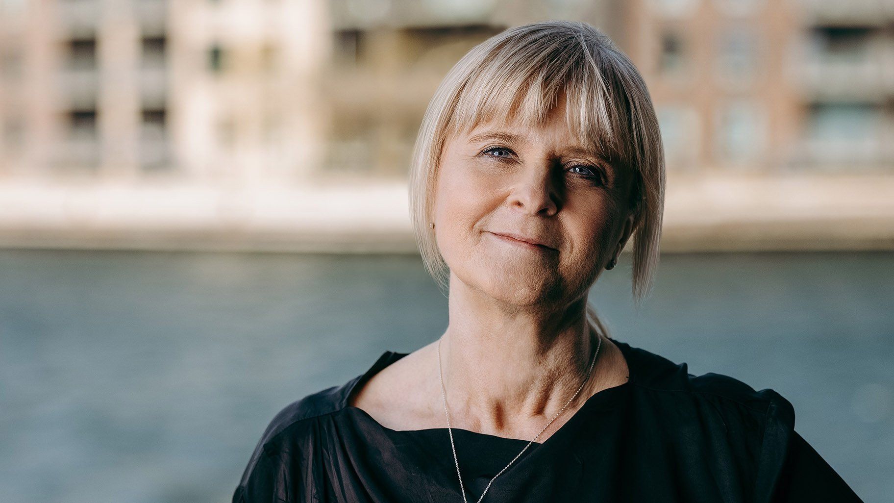 Kvinde med blond hår, smilende, udendørs nær vand, iført en sort top.