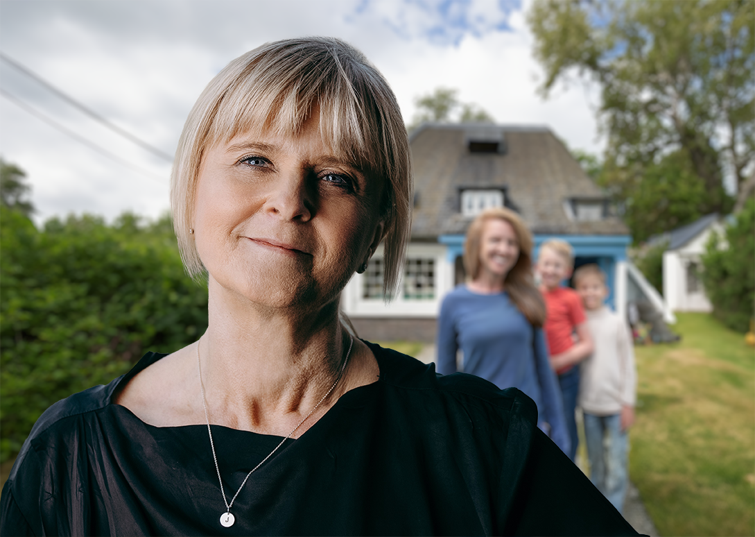 Smilende kvinde i forgrunden med et hus og en familie i den slørede baggrund.