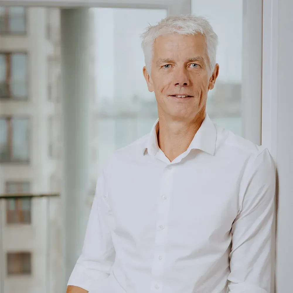 Mand med gråt hår iført en hvid skjorte med knapper, lænet op ad en væg indendørs, smilende let.
