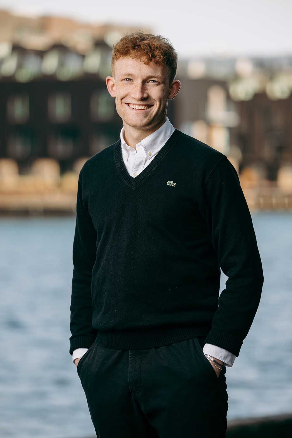 Mand med rødt hår, smilende, iført en sort sweater over en hvid skjorte, stående udenfor med et vandfald i baggrunden.