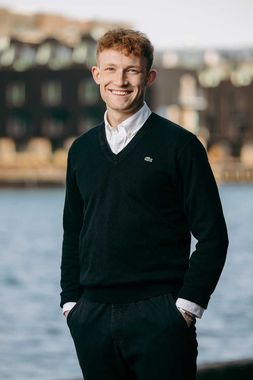 Mand med rødt hår, smilende, iført en sort sweater over en hvid skjorte, stående udenfor med et vandfald i baggrunden.