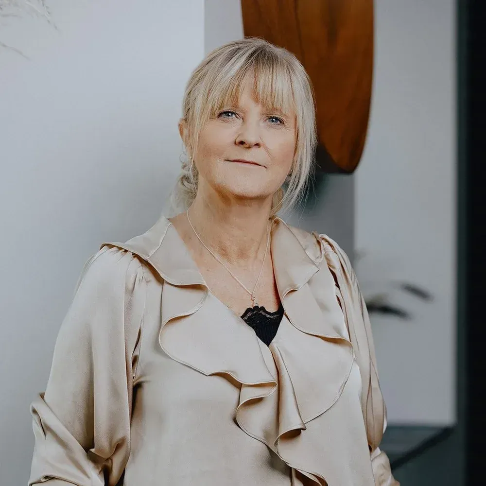Kvinde med blond hår, iført en beige jakke, stående indendørs.