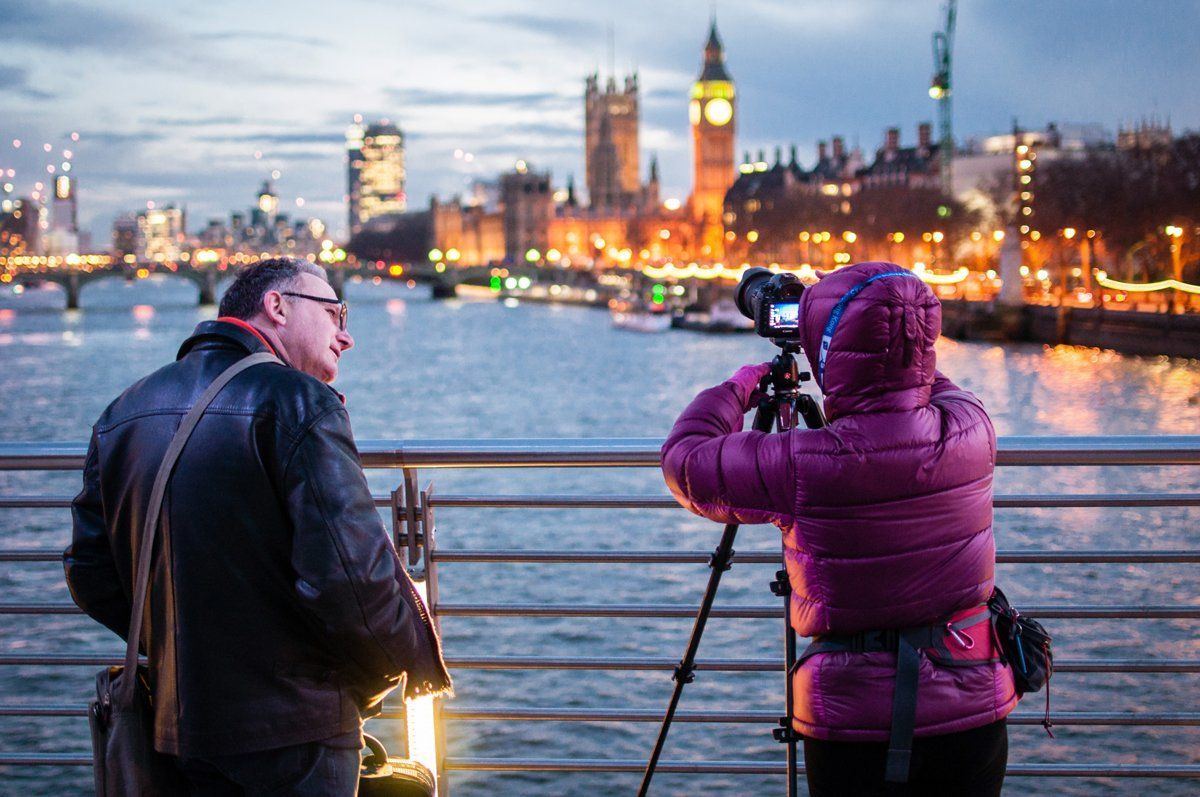 To personer fotograferer Londons skyline i skumringen. En person bruger et stativ. Flod og lys i baggrunden.