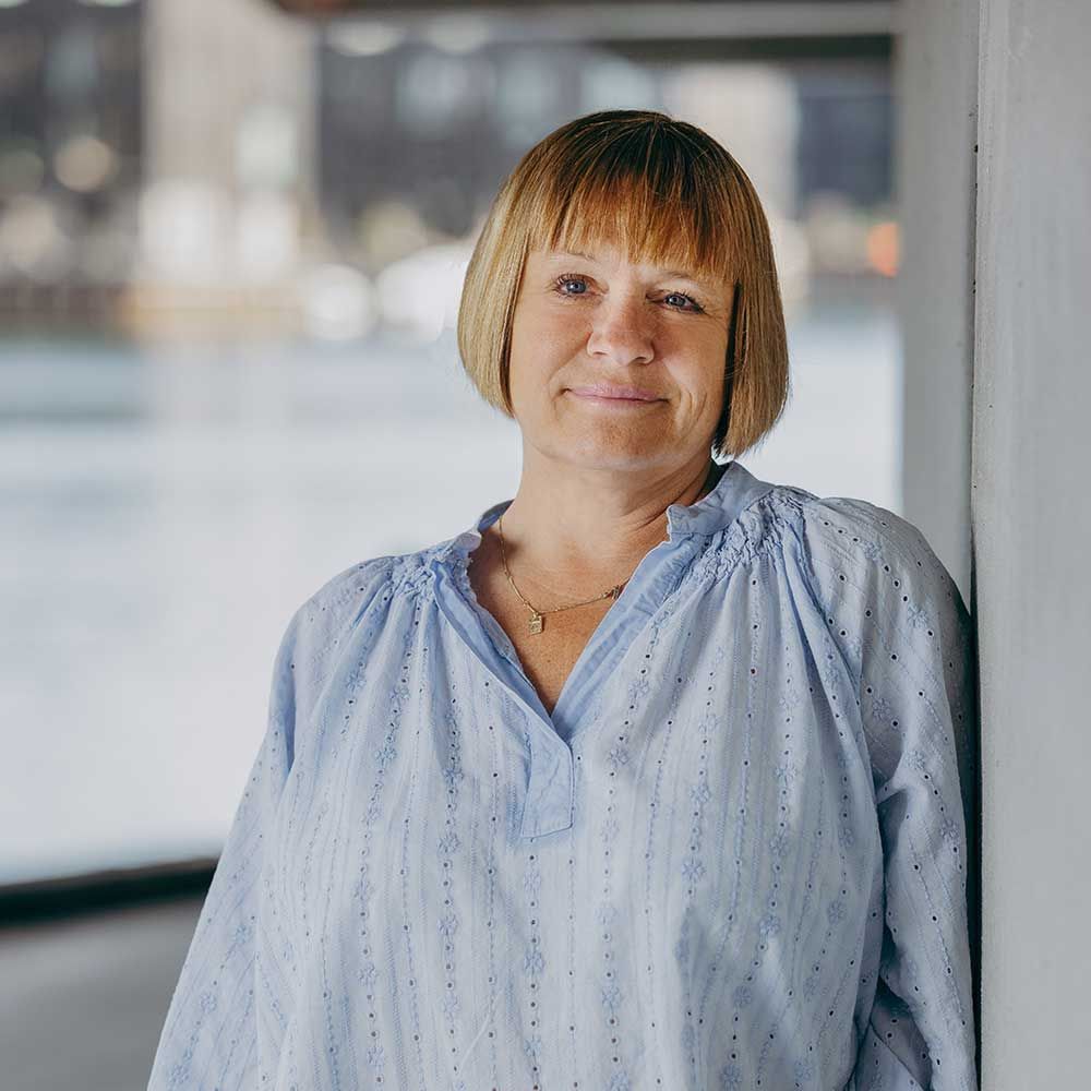 Kvinde med kort blond hår i en blå top læner sig smilende op ad en hvid væg udendørs.