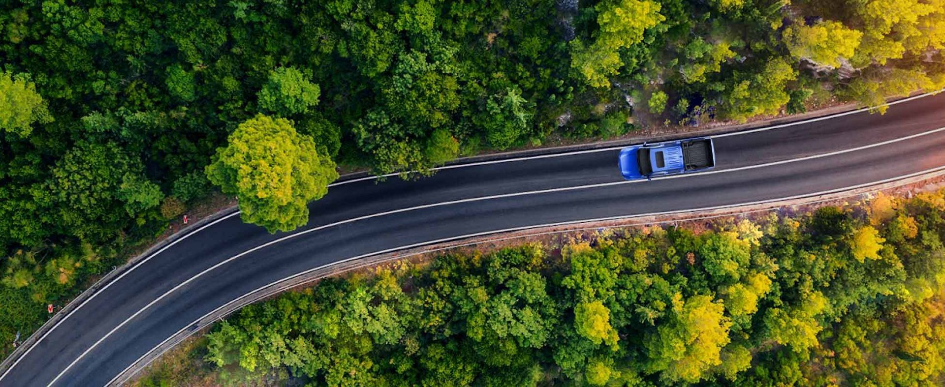 Vista aérea de una sinuosa carretera de montaña con dos coches atravesando un denso bosque verde.