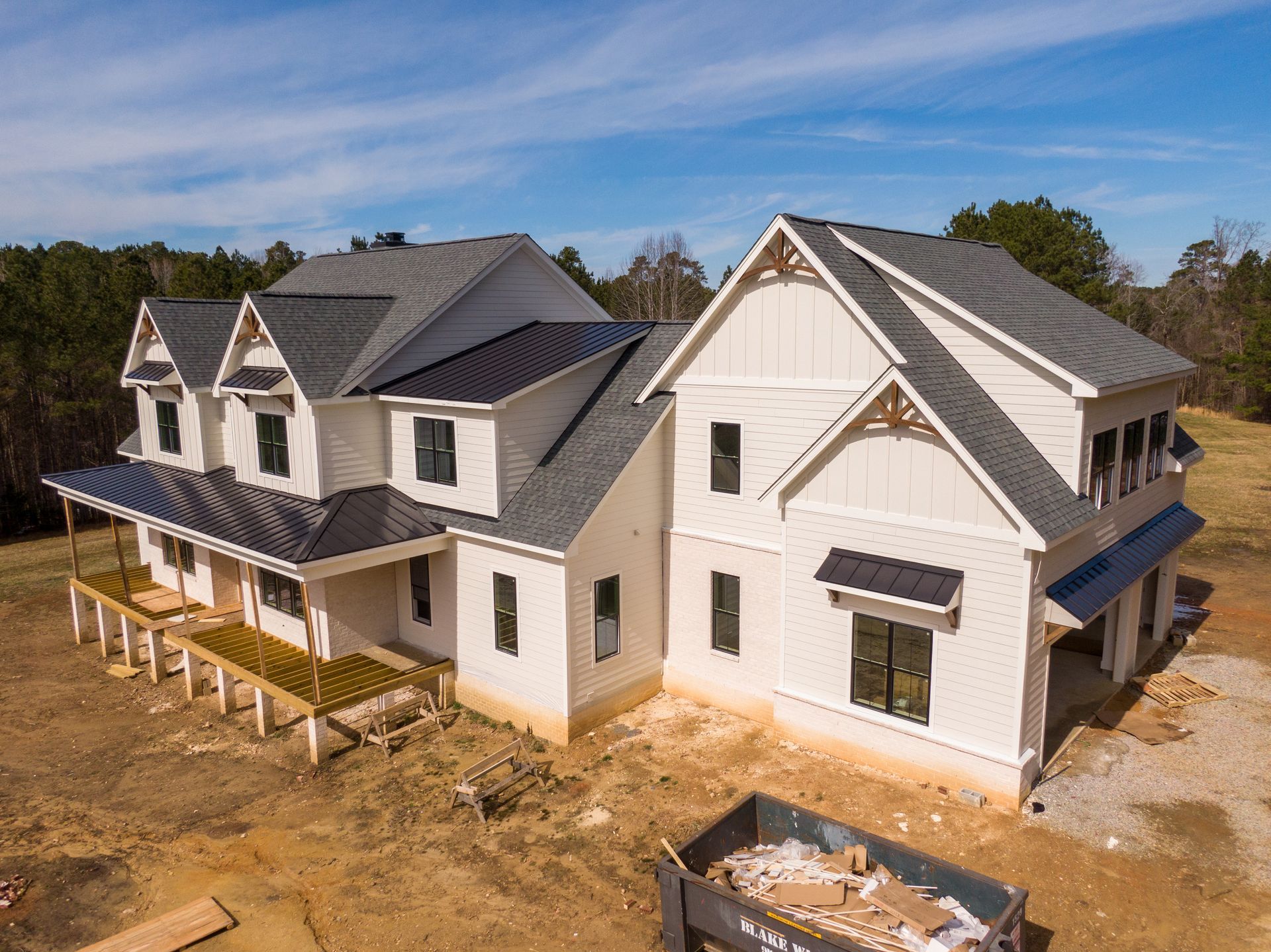 A newly constructed two-story white farmhouse with a dark metal and shingle roof, set on a dirt lot with a dumpster. A newly constructed two-story white farmhouse with a dark metal and shingle roof, set on a dirt lot with a dumpster.